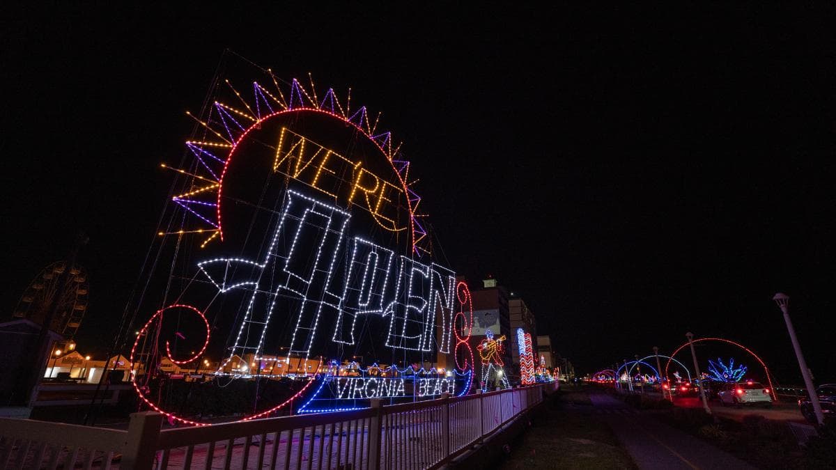 Colorful holiday light display at night with "We're Open Virginia Beach" in bright letters along a boardwalk.