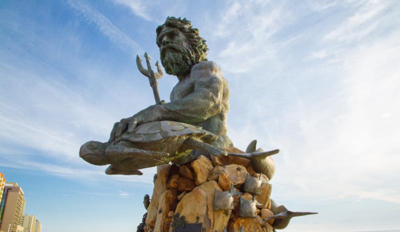 Bronze statue of King Neptune holding a trident, seated on a rock with marine life sculptures, under a blue sky.