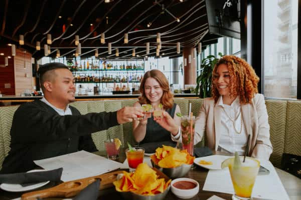 Three people sitting at a restaurant table clinking glasses, with drinks, chips, and menus in front of them.