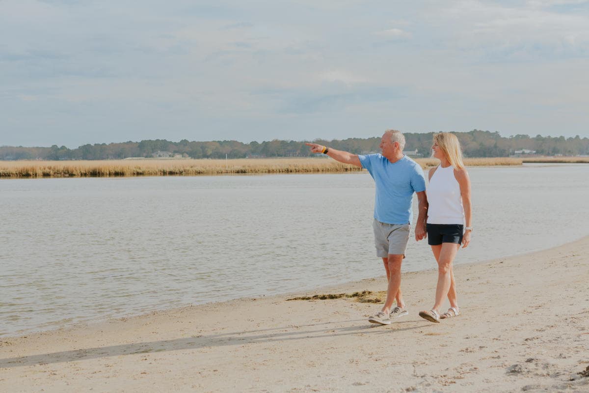 An older couple walks hand in hand along a sandy beach. The man is pointing at something in the distance.