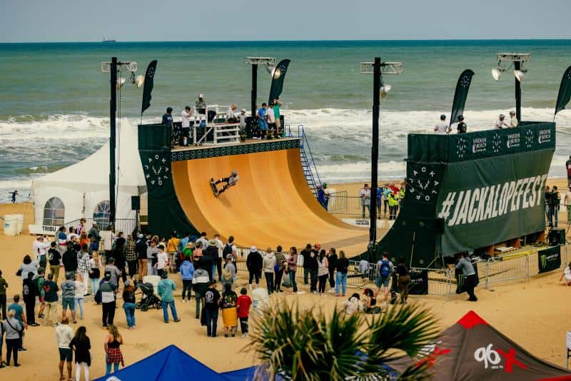 A crowd watches a skateboarder perform on a large ramp at the Jackalope Festival, with ocean waves in the background.