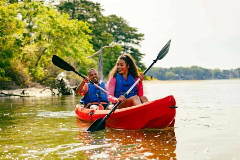 Two people in life jackets kayaking on a calm lake, surrounded by trees and clear skies.