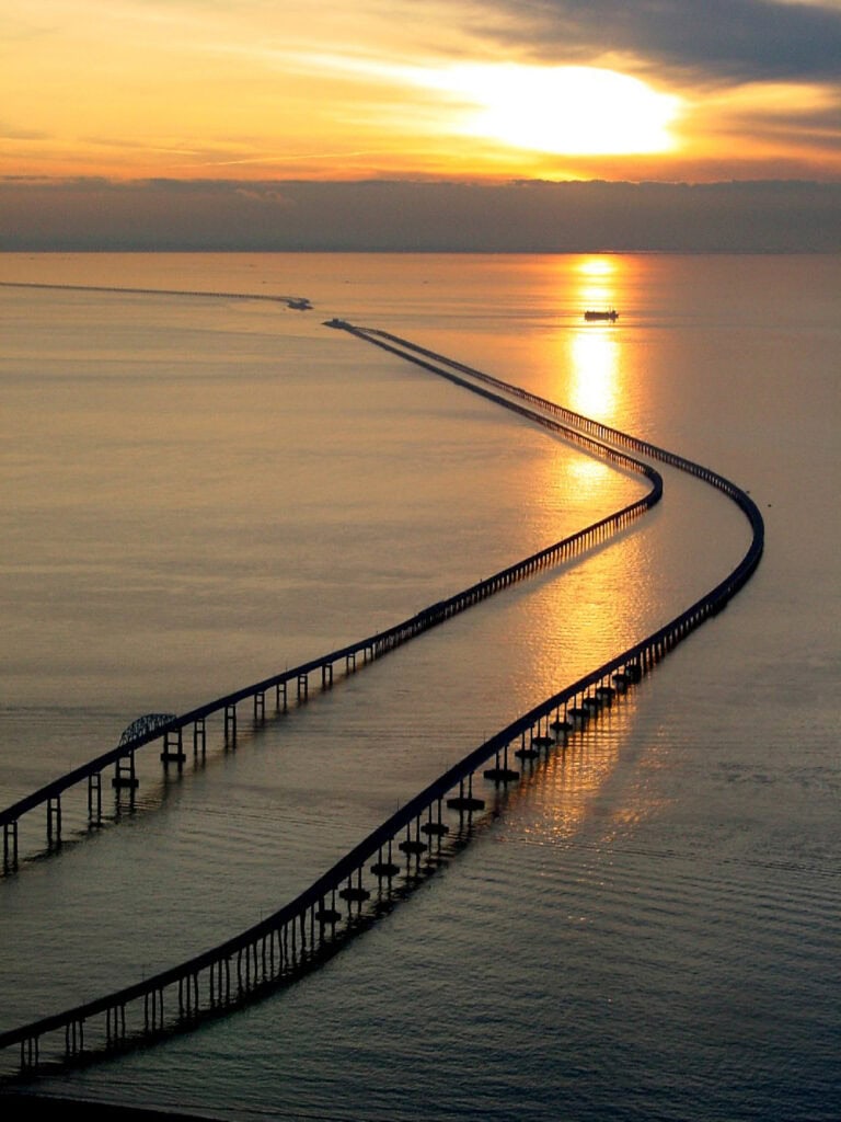 Aerial view of a curved bridge extending over calm waters during sunset, reflecting golden hues in the sky.