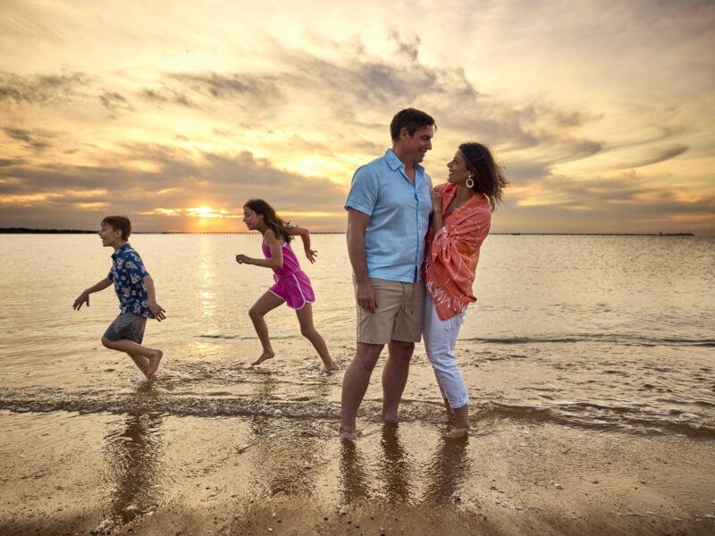 A couple embraces on the beach at sunset, while two children playfully run through the water, creating a joyful family moment.
