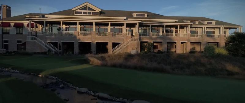 Sunlit exterior of a large, two-story building with a green lawn and rocky stream in the foreground.
