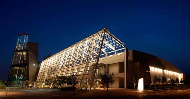 Modern building with an illuminated glass facade and angular design, set against a dark blue evening sky. Trees and a lit pathway are visible in the foreground.
