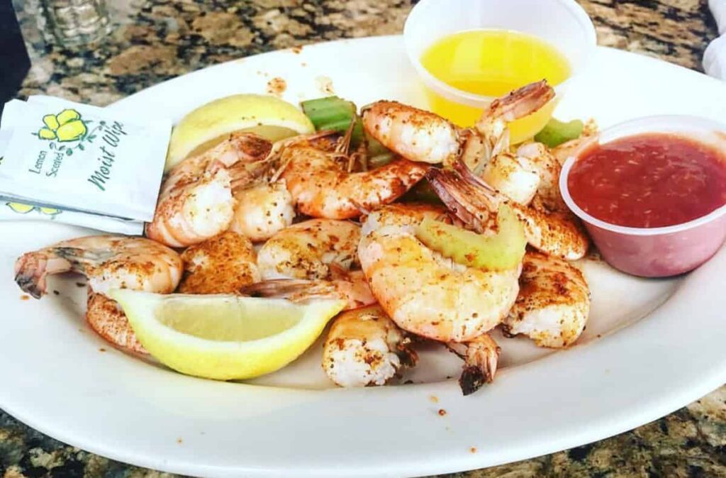 A plate of seasoned shrimp with lemon wedges, a cup of melted butter, cocktail sauce, and a packet of moist towelettes on a granite surface.