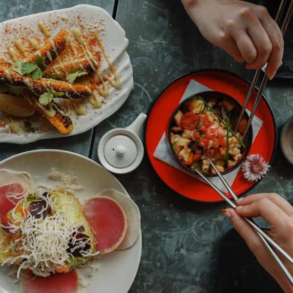 A table spread with a plate of grilled toast, a dish of sliced vegetables with shredded cheese, and a small bowl of mixed salad. Two hands holding chopsticks are reaching for the salad.