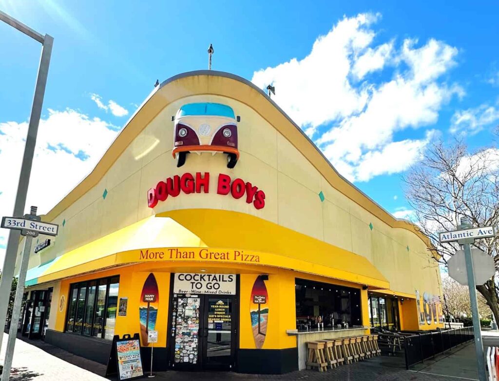 Exterior of Dough Boys restaurant with a unique, curved yellow facade featuring a vintage van design. Signs show "More Than Great Pizza" and "Cocktails to Go." Located at 33rd Street and Atlantic Ave.