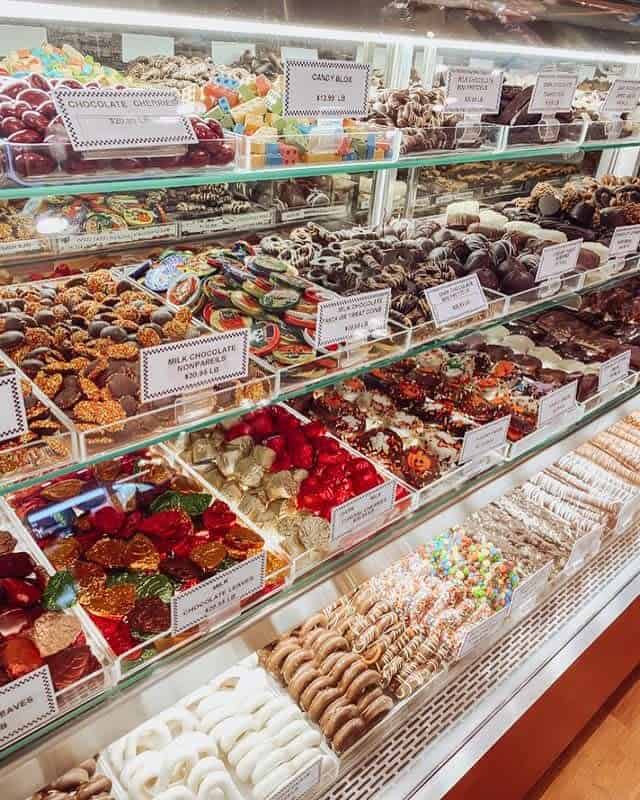 A glass display case filled with various chocolates and candies, including chocolate-covered pretzels, gummies, and nougat, labeled with prices.