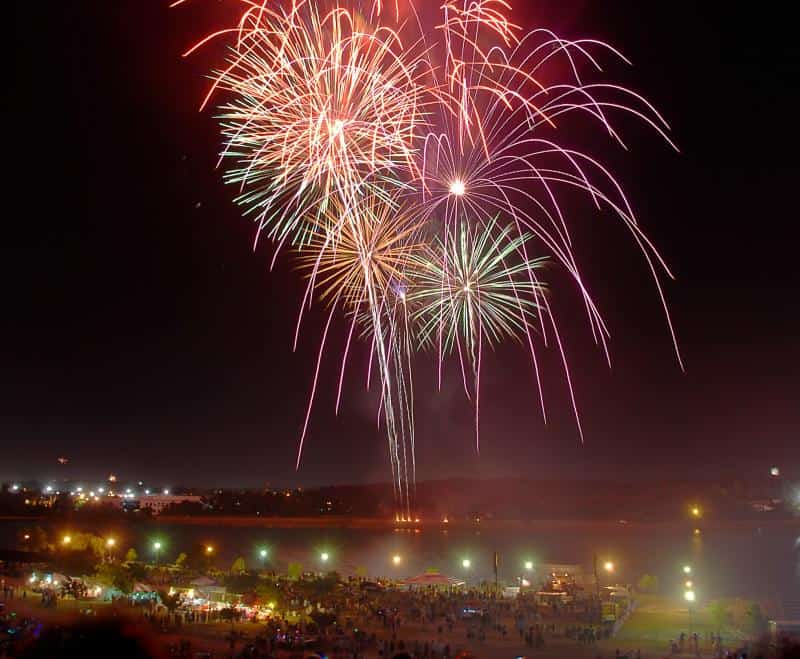 Colorful fireworks burst in the night sky over a cityscape with lights. A crowd gathers below, near a waterfront.