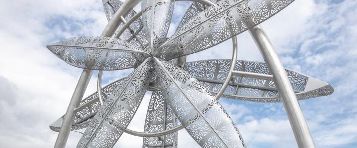 A metallic sculpture with intricate leaf patterns stands against a cloudy sky backdrop.