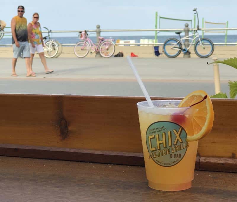 A cocktail in a plastic cup with a lemon slice, labeled "Chix Seaside Grille," sits on a wooden table. In the background, a boardwalk with people walking and bicycles nearby.