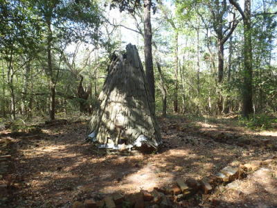 A small, weathered wooden teepee-like structure stands in a wooded area, surrounded by trees and scattered bricks.