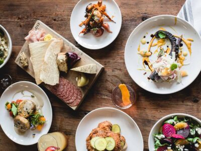 Aerial view of a wooden table with various dishes, including salads, charcuterie, bread, and drinks, arranged in a casual dining setting.
