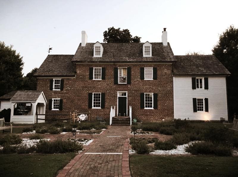 Historic brick and white colonial house with dark shutters, a central pathway, and small front garden, set against a backdrop of trees and an overcast sky.