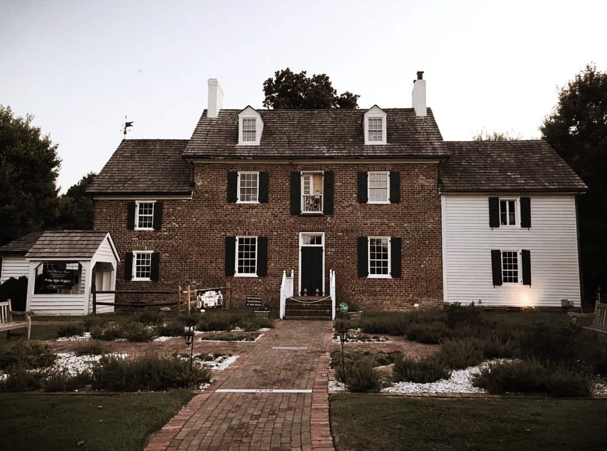 Brick and white colonial-style house with a symmetrical facade, black shutters, and a garden path leading to the entrance. Trees are in the background.
