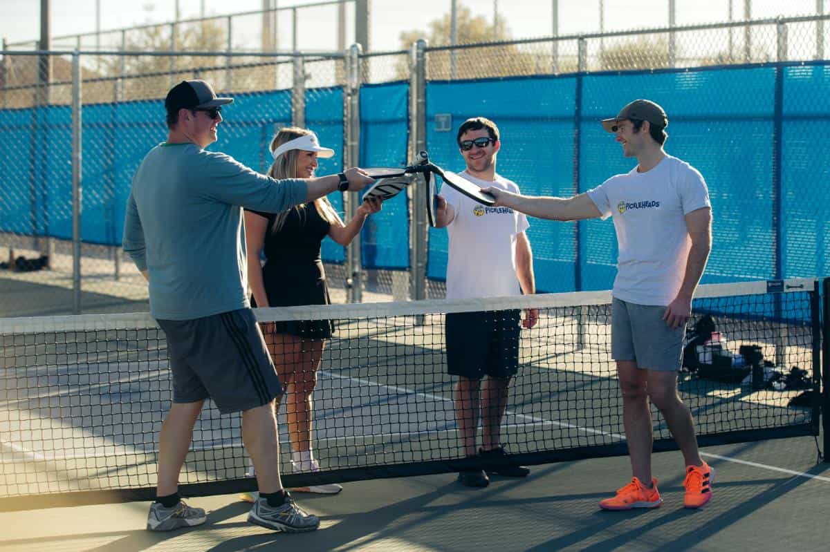 Four people stand on a pickleball court, holding their paddles together over the net, smiling.