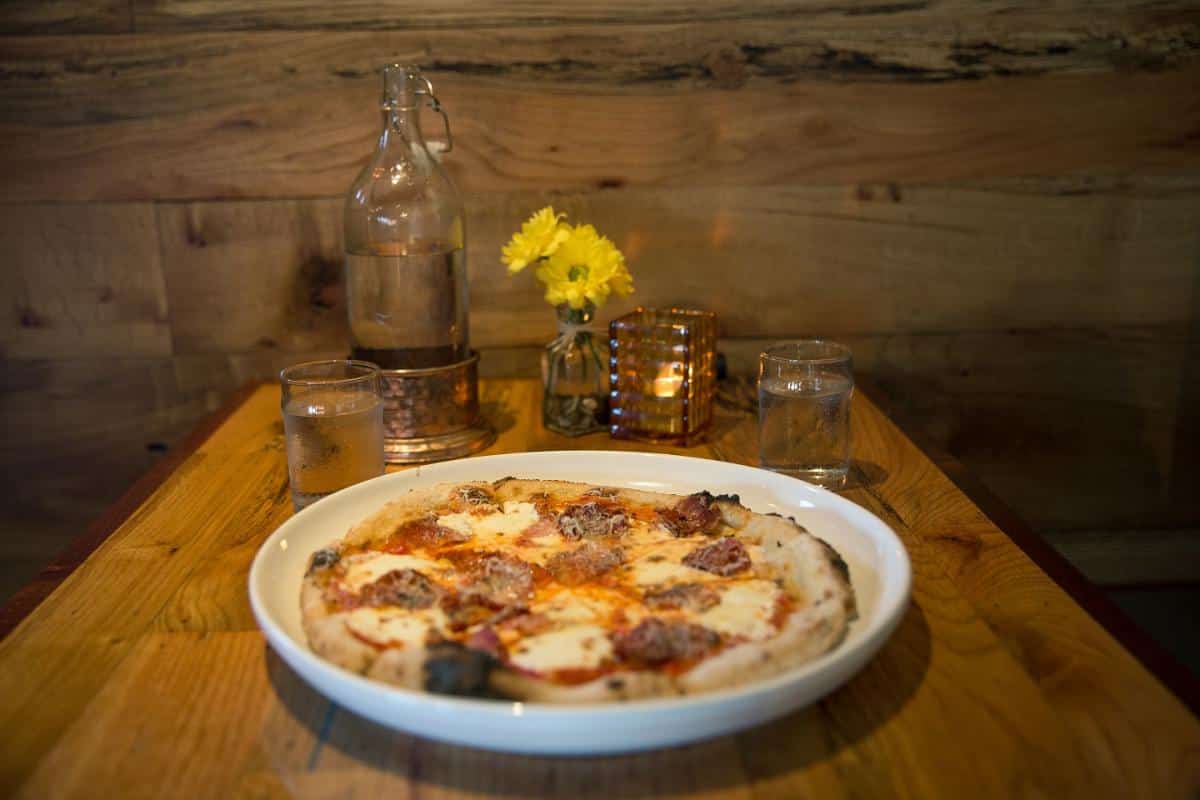 A pizza with sausage and cheese on a wooden table, accompanied by two glasses, a water bottle, yellow flowers, and a candle.