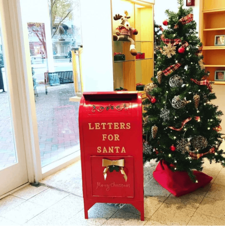 A red "Letters for Santa" mailbox is beside a decorated Christmas tree indoors, near large windows.