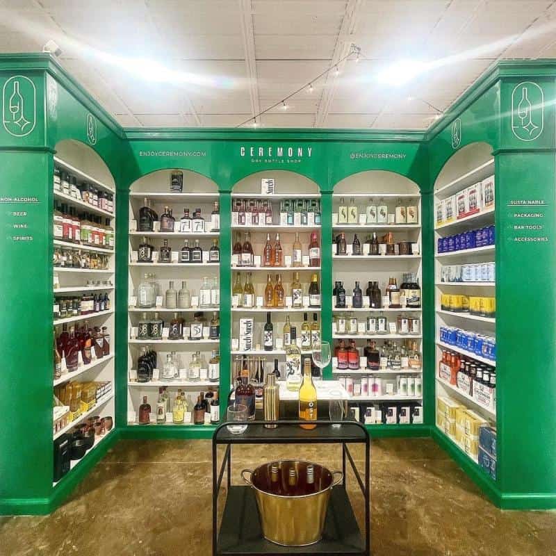 A green-walled retail store display features various bottles and cans of non-alcoholic beverages. A black table with a bucket and bottles is in the center.
