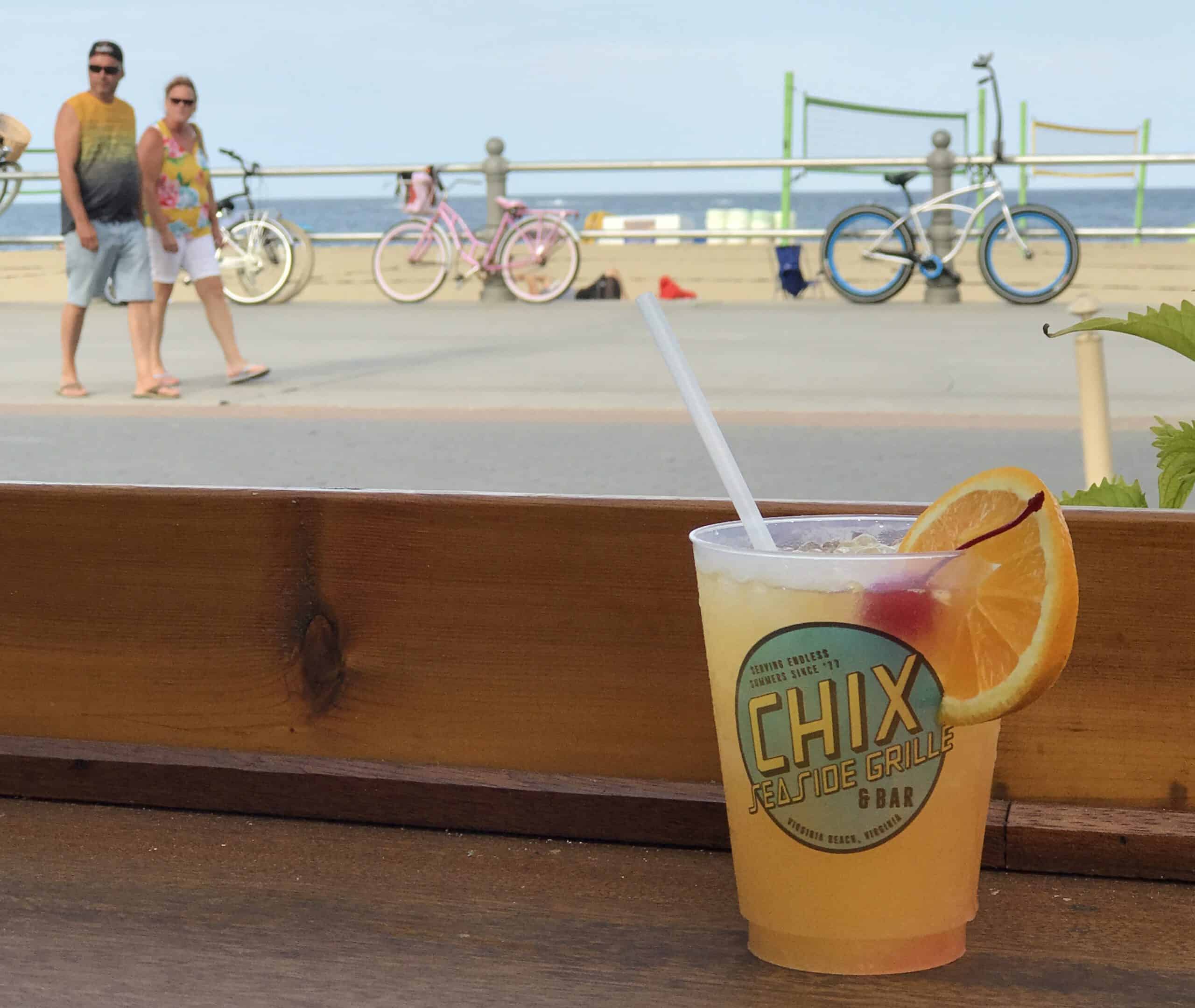 A cocktail with an orange slice sits on a table at Chix Seaside Grille & Bar, with beachgoers and bicycles in the background.