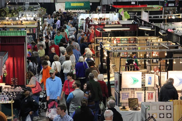 A bustling indoor market scene with crowds of people browsing various vendor booths, featuring art displays and handmade crafts.