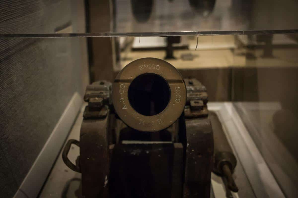 Close-up of a vintage cannon's muzzle under glass. Inscriptions include "A.O. CO," "N4492," "1900," and "H D B." The cannon is mounted on a metal base.