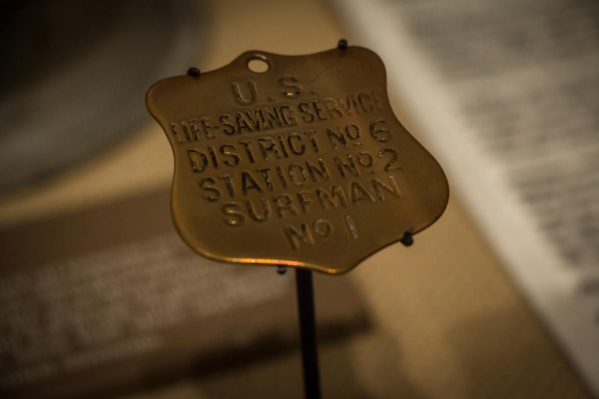 A metal badge reads: "U.S. Life-Saving Service, District No. 6, Station No. 2, Surfman No. 1," displayed on a stand.