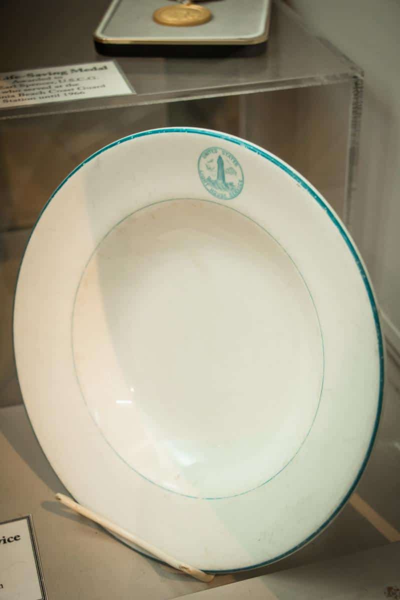 A white porcelain plate with a green border and an emblem, displayed in a glass case. A museum label is visible in the background.