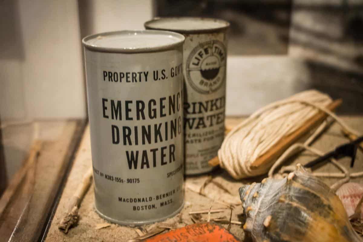 Two cans labeled "Emergency Drinking Water" and "Life Time Drinking Water" are displayed alongside coiled rope and a shell.