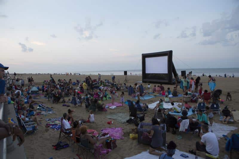 People sitting on a beach with blankets and chairs, watching a movie on a large outdoor screen.