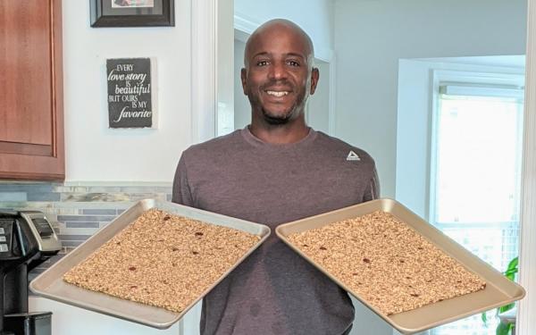 Person standing in a kitchen holding two baking trays with granola. A sign on the wall reads "Every love story is beautiful but ours is my favorite.
