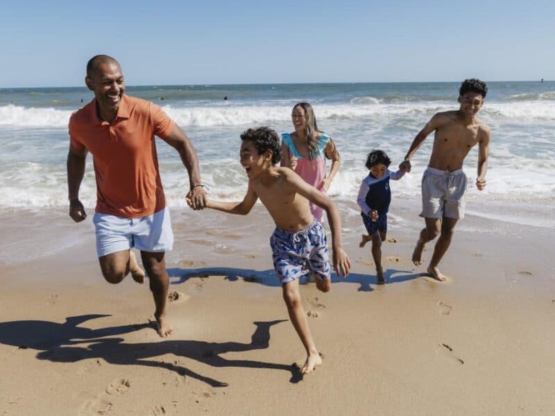 Family enjoying a beach day, running in the sand by the ocean waves under a clear blue sky.