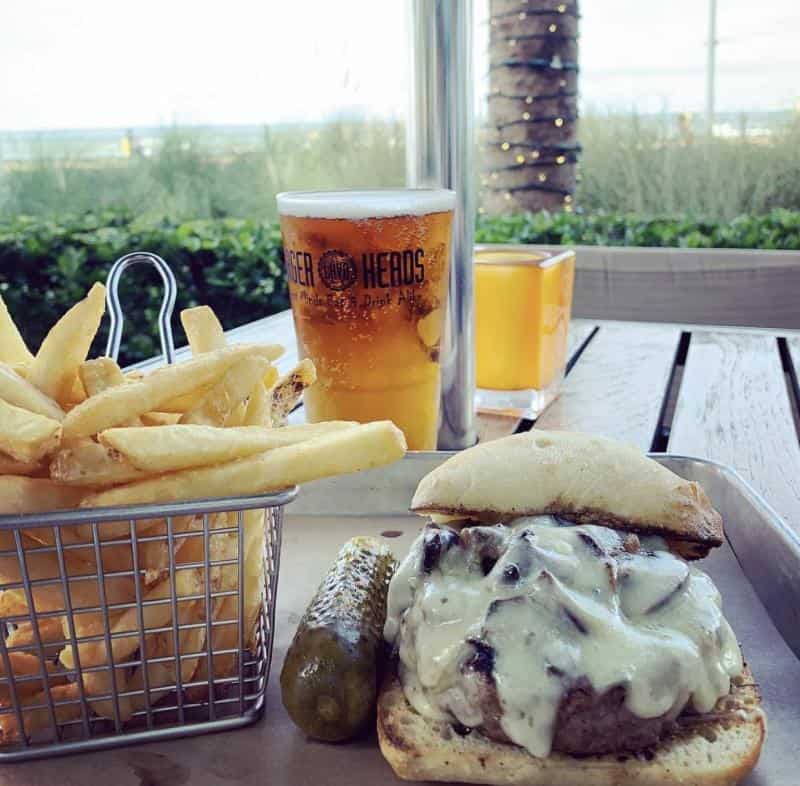 Burger with melted cheese, fries in a basket, pickle, and two beverages on a tray. Outdoor setting with greenery and a view of the ocean in the background.
