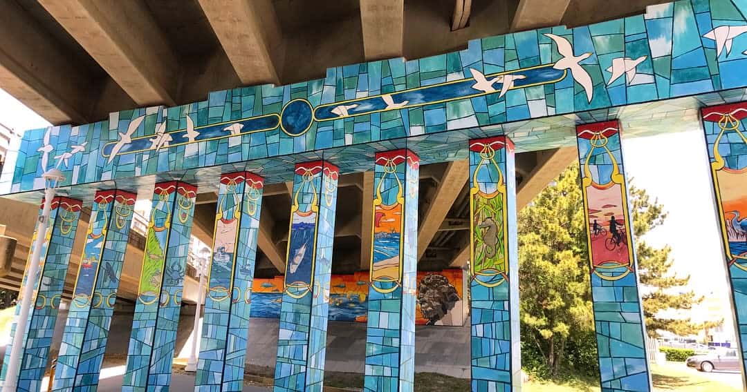 Colorful mural on structural columns under a bridge, featuring abstract designs, birds, and bicycles on a blue and teal background.