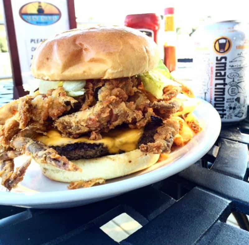 Cheeseburger topped with fried soft-shell crab on a white plate. In the background, there is a soda can, mustard, and ketchup bottles on a black metal table.