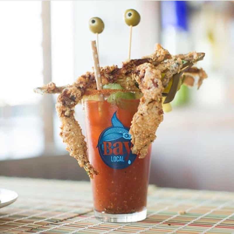 A Bloody Mary cocktail in a glass with "Bay Local" logo, topped with fried foods, two olives on skewers, and lime slices.