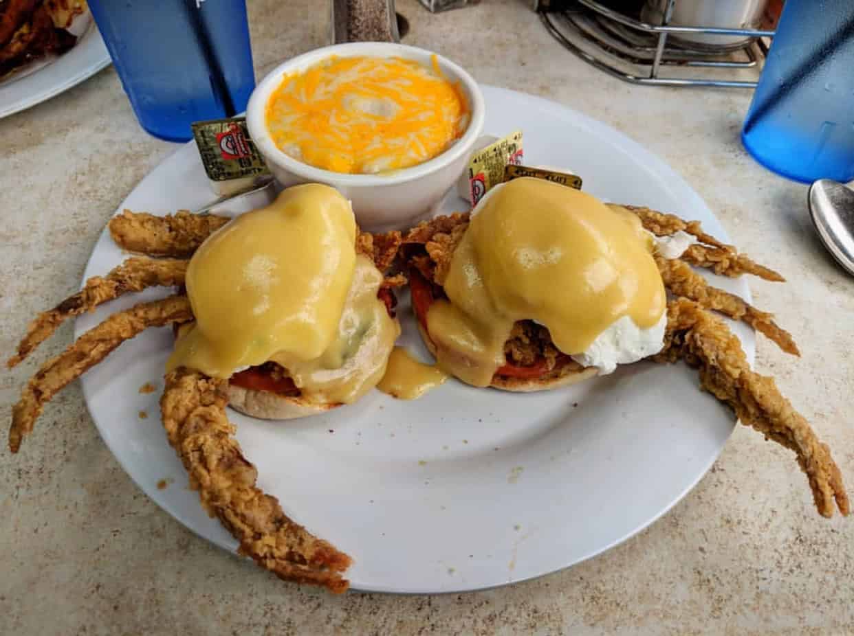 A plate with two soft-shell crab benedicts topped with hollandaise sauce, accompanied by a bowl of shredded cheese and grits, next to blue glass tumblers.