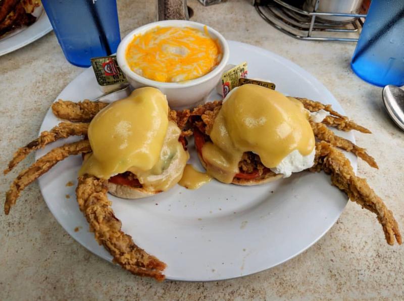 Two fried soft-shell crabs with sauce on English muffins, topped with poached eggs, served with a bowl of cheese grits.