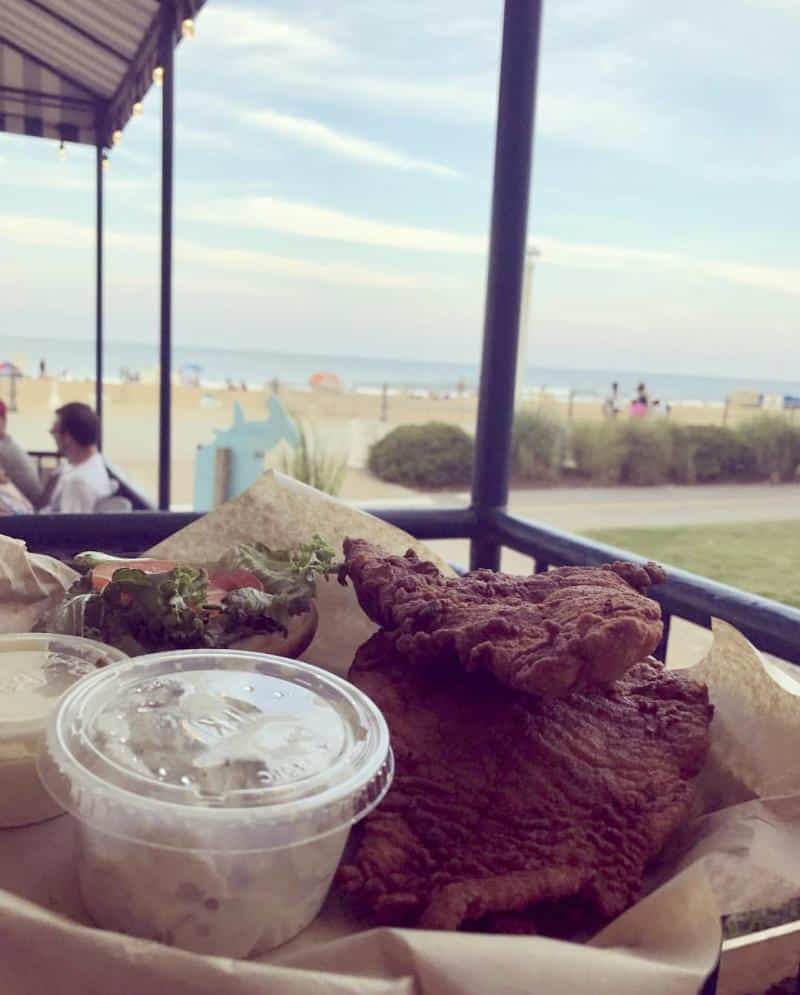Fried fish with a side of coleslaw and salad in a basket, overlooking a sandy beach and ocean view from a covered outdoor seating area.