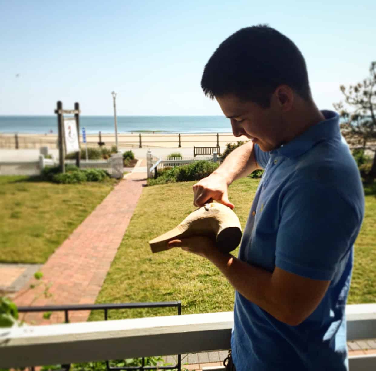 Man carving a piece of wood on a porch with a beach and ocean in the background.