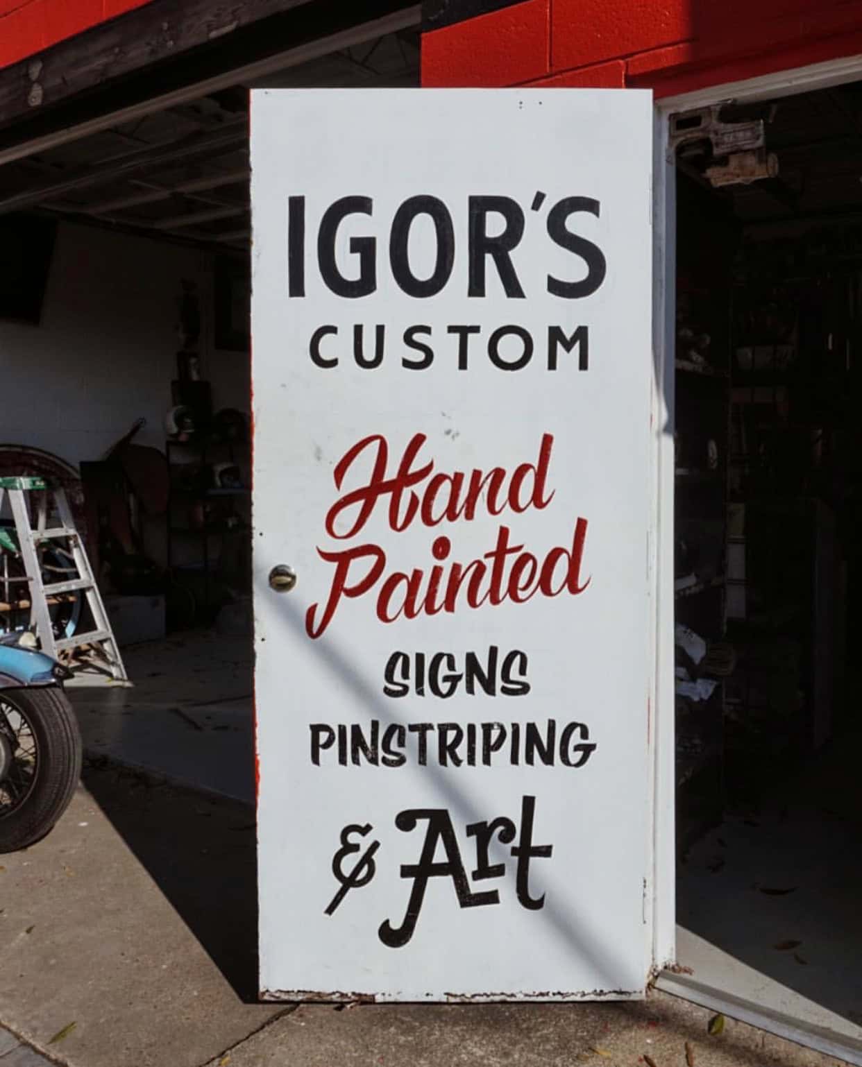 A door displays the text: "Igor's Custom Hand Painted Signs Pinstriping & Art," situated in front of a garage with a ladder and tools visible inside.