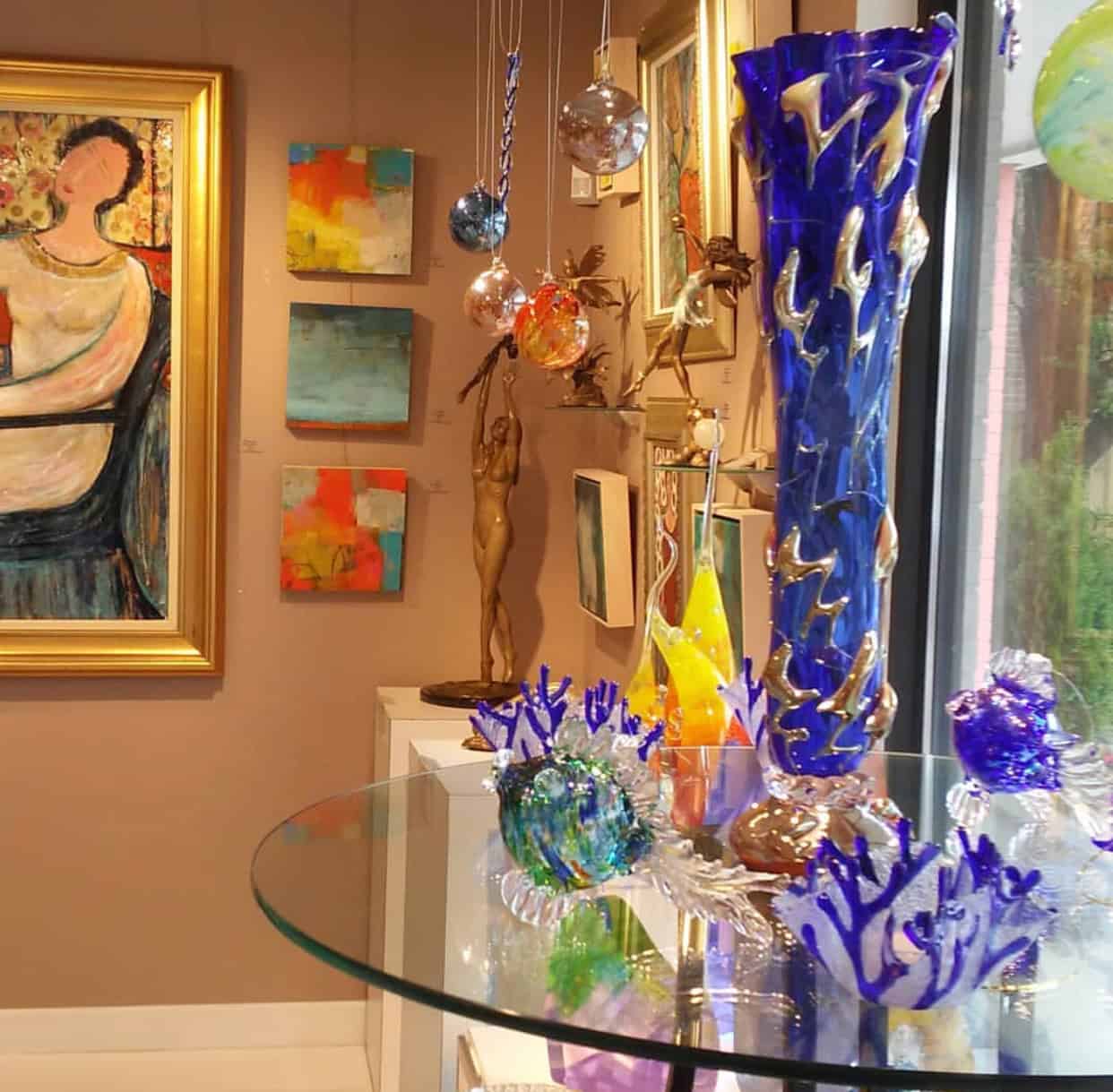 Art gallery display with abstract paintings on the walls, a large blue glass sculpture, and smaller colorful glass pieces on a round glass table.