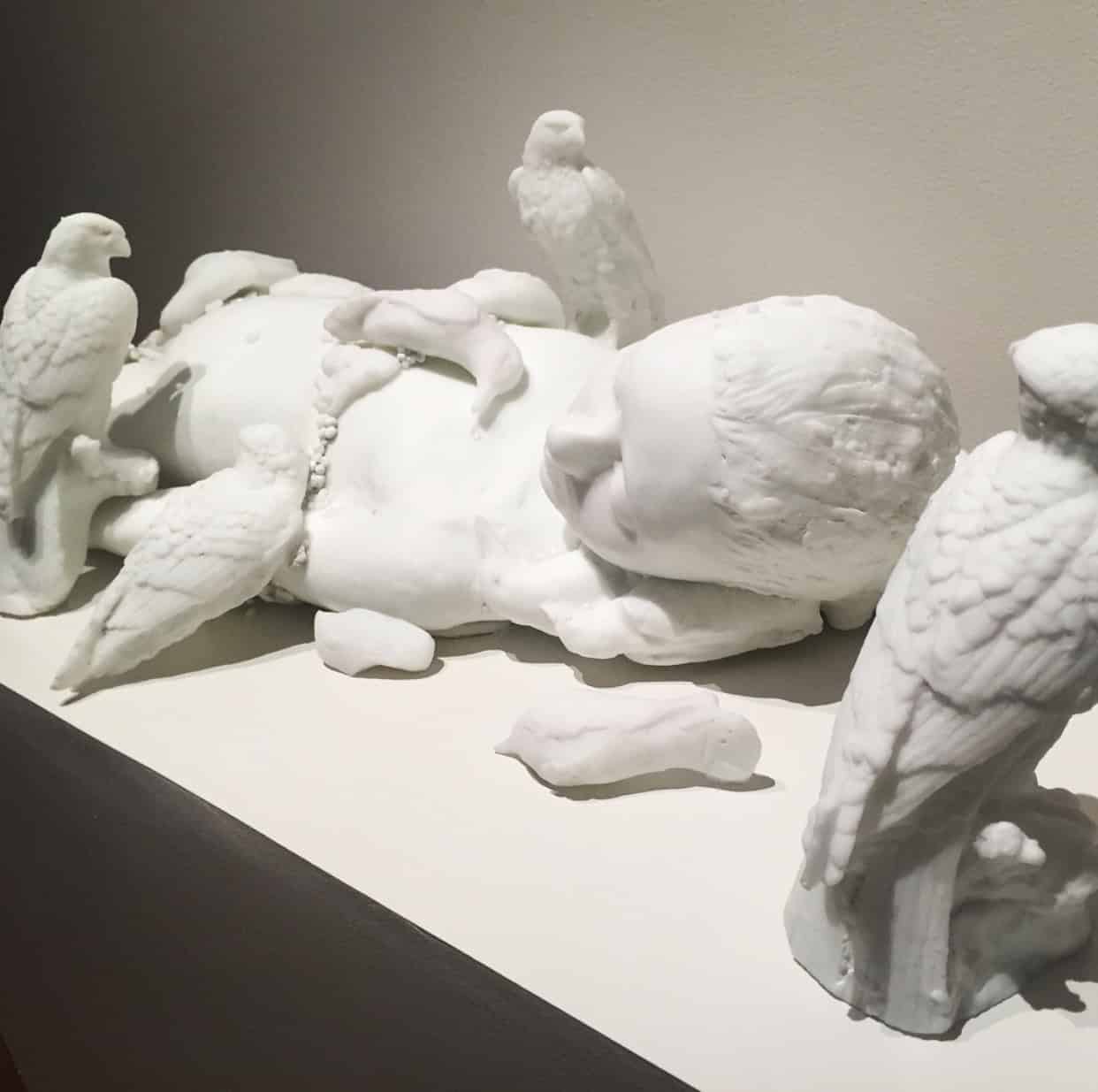 A white sculpture of a reclining human figure surrounded by several birds on a flat surface.