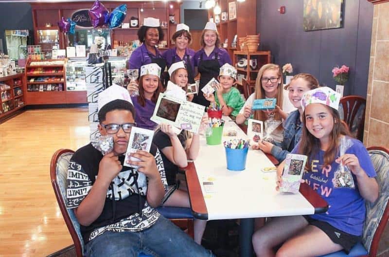A group of children sitting at a table in a shop, wearing paper hats and holding craft items, with three adults standing behind them. Balloons and shelves of products are visible in the background.