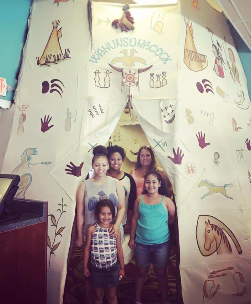 Five people standing in front of a decorated teepee entrance with various symbols and illustrations.