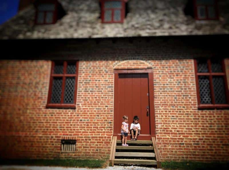Two children sit on wooden steps in front of a brick building with a red door and windows.