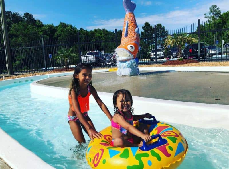 Two children in swimsuits play in a shallow pool. One child sits on a colorful inflatable ring, while the other stands nearby. A large fish statue is in the background.