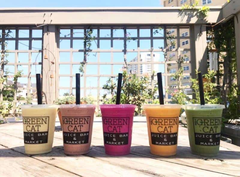Five colorful smoothies in clear cups with black straws, labeled "Green Cat Juice Bar & Market," lined up on a wooden table with a cityscape background.
